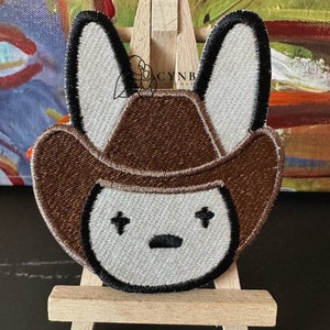 May include: Embroidered patch of a cartoon bunny wearing a brown cowboy hat with white ears and a white face. The bunny has black eyes and a black mouth.