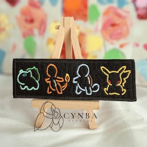 May include: A rectangular black embroidered patch featuring four outlined character figures in light green, orange, light blue, and yellow thread. The patch is displayed on a small wooden easel. The logo for Cynba Studios is visible below.