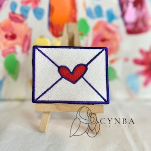 May include: A white envelope with a purple border and a red heart in the center. The envelope is displayed on a small wooden easel.