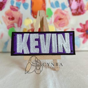 May include: A rectangular patch with the name "KEVIN" embroidered in white on a purple background, framed by a black border. The patch is displayed on a small wooden easel. The logo "CYNBA STUDIOS" is visible below.