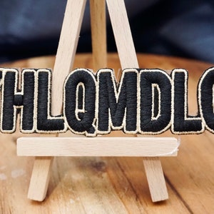 May include: Black embroidered patch with the letters "YHLQMDLG" in white outline. The patch is displayed on a small wooden easel.
