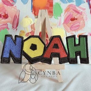 May include: A colorful embroidered patch spelling out the name "NOAH" in block letters. The letters are outlined in black and each letter is a different color: blue, yellow, red, and green.