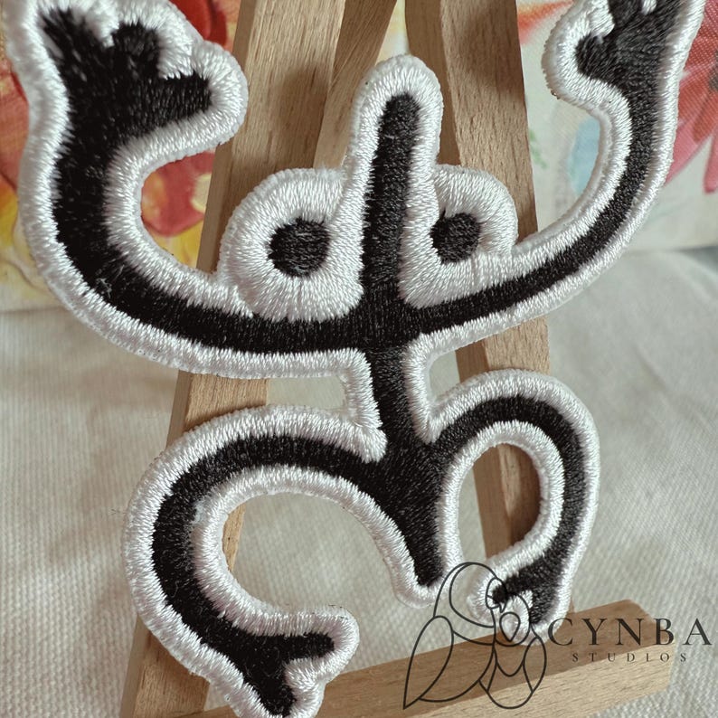 May include: Black and white embroidered patch with a stylized figure. The design features a black figure with white outlining, arms raised, and a unique shape. The patch is likely for clothing or accessories.