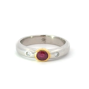 May include: A silver ring with a gold bezel-set oval ruby gemstone in the center. Two small, round, clear gemstones are set on either side of the ruby. The ring has a smooth, polished finish.