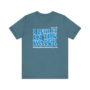 May include: A teal short-sleeve t-shirt with the text "I LEFT IT IN THE WATER" in a blue, bubble-style font. The text "Acts 2:38" is above the main text, and "kaylagabbardministry.com" is below. The shirt is made of a soft material.