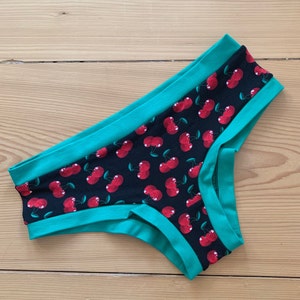 Op de afbeelding: Zwarte onderbroek met een patroon van rode kersen. De randen zijn afgezet met een felgroene stof. Het ondergoed is gemaakt van een zacht, comfortabel materiaal.