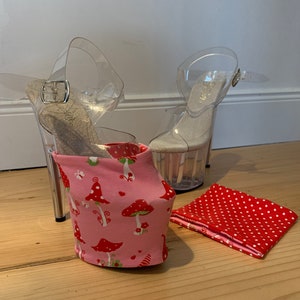 Può includere: Sandali con tacco alto e plateau trasparenti con cinturini alla caviglia e una fodera in tessuto rosa con funghi e cuori rossi. Un piccolo quadrato di tessuto rosso a pois è accanto alla scarpa. Le scarpe sono su una superficie di legno.