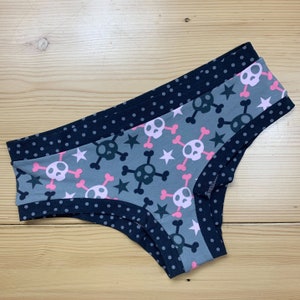 Può includere: Intimo grigio con un motivo di teschi e ossa incrociate, accenti rosa e stelle. I bordi sono rifiniti con tessuto blu navy a pois bianchi. L'intimo è realizzato in un materiale morbido e confortevole.