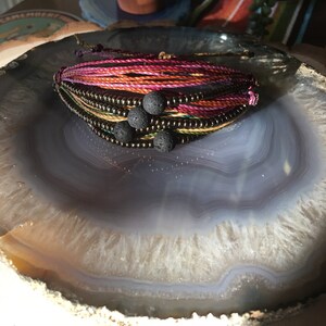May include: A multi-colored string bracelet with black lava beads. The bracelet is on a gray and white agate slice.