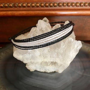 May include: Black and white friendship bracelet with multiple strands of cord, displayed on a white crystal.