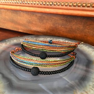 May include: Two bracelets with black beads and lava rock beads. The bracelets are layered on top of each other. The top bracelet is made of multi-colored string. The bottom bracelet is made of black beads and lava rock beads.