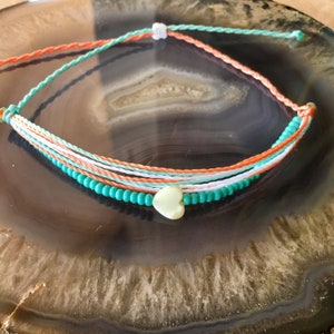 以下が含まれることがあります： ハートのチャームが付いたマルチストランドブレスレット。ブレスレットは白、オレンジ、ターコイズの糸とターコイズビーズで作られています。