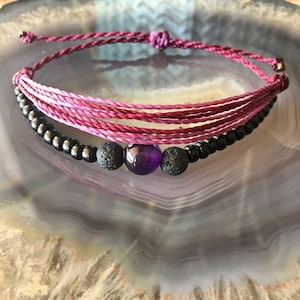 Może przedstawiać: Bransoletka z ametystem w kolorze fioletowym z czarnymi koralikami lawowymi i czarnymi koralikami nasion. Bransoletka wykonana jest z różowo-fioletowego sznurka ombre.