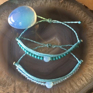 May include: Three turquoise blue bracelets with a light blue opal bead on each. The bracelets are made of string and beads. The bracelets are on a gray and white agate stone background.