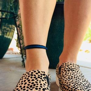 May include: A person wearing a black and blue ankle bracelet with a thin blue stripe. They are wearing leopard print slip-on shoes.