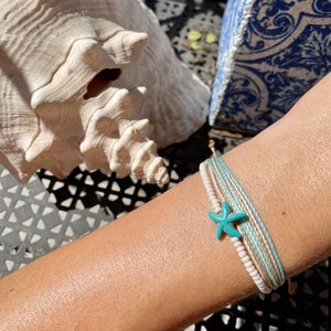 May include: A turquoise starfish charm on a multi-layered bracelet with white, beige, and brown cords.