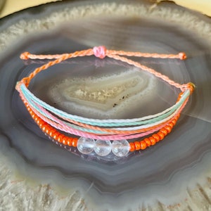 May include: A multi-strand bracelet with orange, pink, and white string, and clear crystal beads.