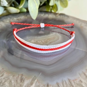 Könnte beinhalten: Ein rotes und weißes Freundschaftsarmband aus Schnur. Das Armband ist verknotet und liegt auf einem grauen und weißen Achat-Stein.