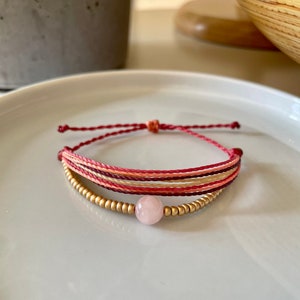 May include: A multi-strand bracelet with pink, red, and beige threads, accented with gold beads and a pink gemstone charm.