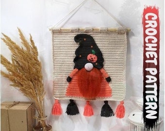 Op de afbeelding: Haakpatroon voor een Halloween-dwerg wandhanger. De dwerg draagt ​​een zwarte hoed met een oranje pompoen en heeft een lange oranje baard. De wandhanger heeft een beige achtergrond en zwarte en oranje kwastjes.