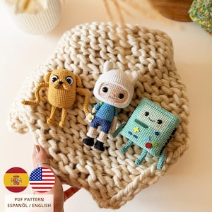 Pode incluir: Três figuras de crochê representando personagens de Adventure Time: um cão amarelo, um humano com gorro branco e um console retangular azul claro. As figuras estão sobre um tecido de malha grossa cor creme. Texto: PDF PATTERN ESPAÑOL / ENGLISH.