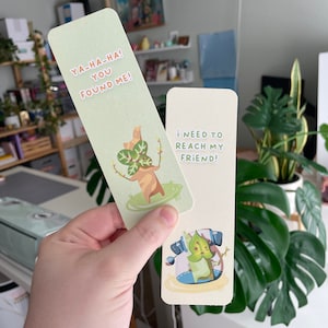 May include: Two illustrated bookmarks. One light green bookmark features a cartoon character with the text "YA-HA-HA! YOU FOUND ME!". The other bookmark is off-white with the text "I NEED TO REACH MY FRIEND!" and a cartoon character.