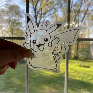 May include: A clear, iridescent sticker of a cartoon Pikachu with a cracked glass design. The sticker is on a window with a blurry background.