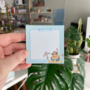 May include: A blue and white gridded notepad with a cartoon mole character holding a pickaxe. The mole is wearing a blue jumpsuit, a yellow hard hat, and a surprised expression. The text "!!!" is above the mole.