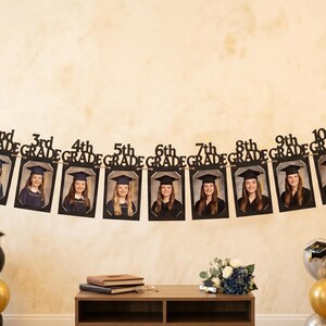 May include: A graduation banner with photos of students from Kindergarten to 12th grade. The banner is black with gold accents and is strung across a wall. Balloons in gold, silver, and black are in the foreground.