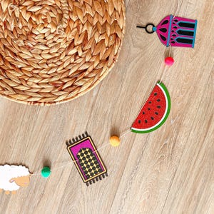 May include: A colorful garland with a watermelon slice, a mosque, a sheep, and a prayer rug. The garland is strung with colorful pom-poms.