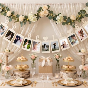 May include: A decorative display featuring a banner with the text BRIDE TO BE, interspersed with hanging photo frames, draped over a sheer curtain adorned with artificial eucalyptus and cream roses, with tiered dessert stands and floral arrangements.