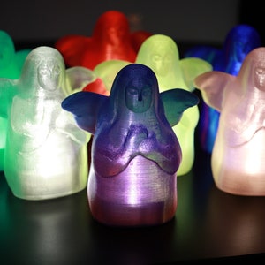 May include: A group of seven small, translucent angel figurines, each with a different color, including green, white, red, yellow, blue, and pink. The angels are all in a praying position with their hands clasped together.