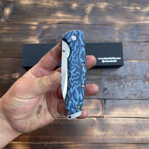 Könnte beinhalten: Ein Klappmesser mit einem blau-schwarz gemusterten Griff wird in einer Hand gehalten. Die Klinge ist silberfarben und teilweise geöffnet. Der Griff hat ein einzigartiges, wirbelndes Design. Das Messer liegt auf einer dunklen Holzoberfläche.