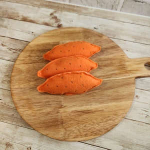 May include: Three orange felt sweet potatoes with black speckles are arranged on a round wooden cutting board. The cutting board has a handle and is placed on a light wood surface.