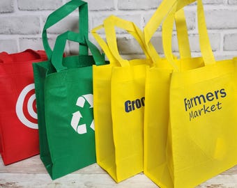 Puede incluir: Un conjunto de cuatro bolsas de la compra reutilizables en rojo, verde y amarillo. La bolsa roja presenta un logotipo de diana circular blanco. La bolsa verde muestra un símbolo de reciclaje blanco. Las bolsas amarillas tienen texto azul Groceries y Farmers Market.