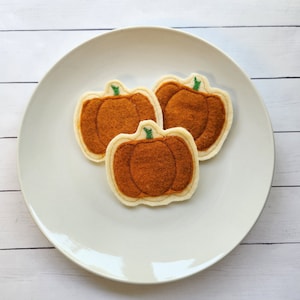 May include: Three felt pumpkin-shaped ornaments on a white plate. The pumpkins are brown with green stems.