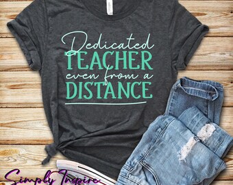 Może przedstawiać: Szary t-shirt z napisem "Dedicated Teacher even from a DISTANCE" w białym i turkusowym kolorze. Koszulka leży na drewnianej powierzchni z niebieskimi dżinsami i białymi butami.