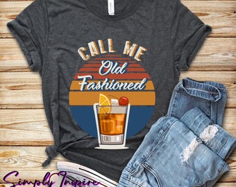 May include: Dark gray t-shirt with the text "CALL ME Old Fashioned" above a graphic of an Old Fashioned cocktail. The design includes a sunset-style background and a cocktail glass with a cherry and orange slice. A pair of blue jeans and sneakers are next to the shirt.
