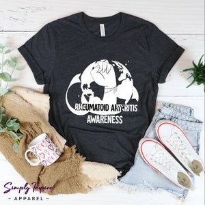 May include: A dark grey t-shirt with a white graphic of a fist holding a globe with the text "Rheumatoid Arthritis Awareness".