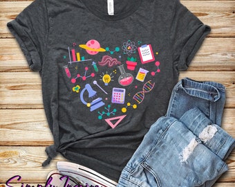 May include: A dark gray t-shirt with a heart-shaped design featuring various science-related icons, including a microscope, a beaker, a calculator, a DNA strand, and a light bulb. The icons are in a variety of colors, including pink, blue, yellow, and green.