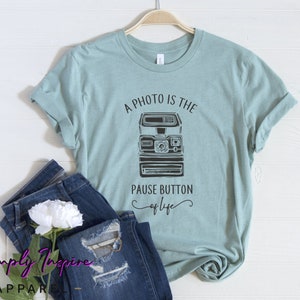 May include: A light blue t-shirt with a black and white graphic of an instant camera and the text "A photo is the pause button of life".