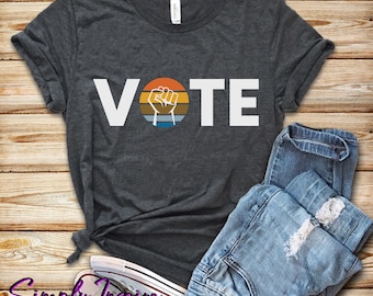 May include: A dark grey t-shirt with the word "VOTE" in white letters. A raised fist with a retro sunset design is in the middle of the word "VOTE".