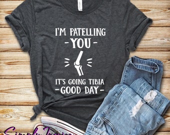 May include: A dark gray t-shirt with the text "I'm patelling -you- It's going tibia -good day-" and a white illustration of a knee bone.