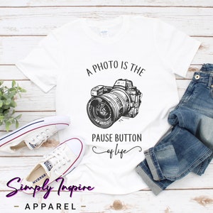 May include: White t-shirt with a black and white illustration of a camera and the text "A photo is the pause button of life".
