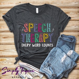 Op de afbeelding: Een donkergrijs T-shirt met de tekst "SPEECH THERAPY EVERY WORD COUNTS" in kleurrijke letters. Het shirt is gevouwen en wordt getoond op een houten oppervlak met een blauwe jeans en witte sneakers.