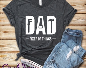 May include: A dark gray t-shirt with the word "DAD" in white letters. The letters are decorated with images of tools, including a wrench, saw, and hammer. The text "FIXER OF THINGS" is printed below the word "DAD".