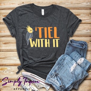 Puede incluir: Una camiseta gris oscuro con un gr&aacute;fico de un p&aacute;jaro cacat&uacute;a amarillo y gris y el texto "'TIEL WITH IT" en letras naranja y amarillas.