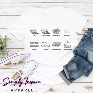 Puede incluir: Camiseta blanca con un gráfico negro de diferentes tipos de nubes y sus nombres: Cirrus, Cirruscumulus, Altocumulus, Altostratus, Stratus, Cumulus, Stratuscumulus, Cumulonimbus. La camiseta está sobre una superficie de madera con un par de zapatillas blancas y unos vaqueros azules.