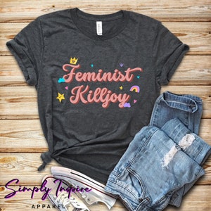 May include: A dark gray t-shirt with the text "Feminist Killjoy" in pink lettering. The text is surrounded by colorful cartoon elements, including a crown, stars, hearts, and a rainbow.