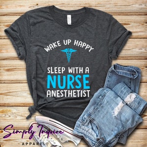 May include: A dark gray t-shirt with the text "Wake Up Happy" above a medical caduceus symbol and the text "Sleep With A Nurse Anesthetist" in blue.